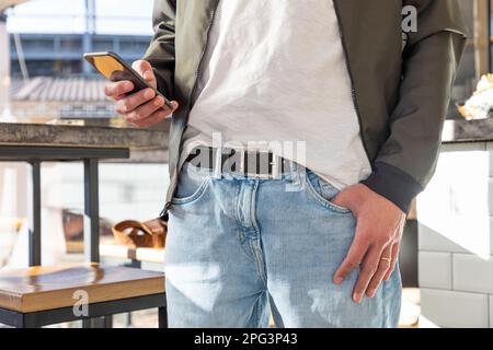 Online-Anwendungen für Smartphones und Werbung. Mann in Jacke und Jeans steht drinnen und benutzt Handy. Nahaufnahme von Händen und Telefon. Bus Stockfoto