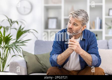 Ein erwachsener, reifer Mann, der allein zu Hause auf dem Sofa sitzt, denkt, er schaut auf das Fenster, deprimiert traurig. Stockfoto