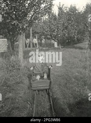 Ein kleiner Junge, der in einem Obstgarten im Vereinigten Königreich auf einem vierrädrigen Holzwagen oder -Wagen (Auto, Kart oder Gokart) auf einer hausgemachten Eisenbahn oder Eisenbahn unterwegs ist. Die Strecke ist ziemlich lang, und das muss eine sehr unterhaltsame Fahrt gewesen sein. In der Ferne beginnt die Strecke auf einer steil abfallenden Rampe, um die Kraft der Schwerkraft zu unterstützen und den Buggy durch den Garten zu fahren. Das ist aus einem schwarz-weißen Glas-Negativ – ein altes, frühviktorianisches/edwardianisches Foto. Stockfoto