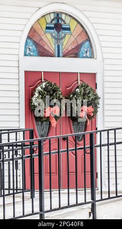 Toivola, Minnesota, USA: Rote Tür der Hope Lutheran Church mit zwei Weihnachtskränzen und einem Regenbogenfenster mit Kreuz an einem Wintertag. Stockfoto