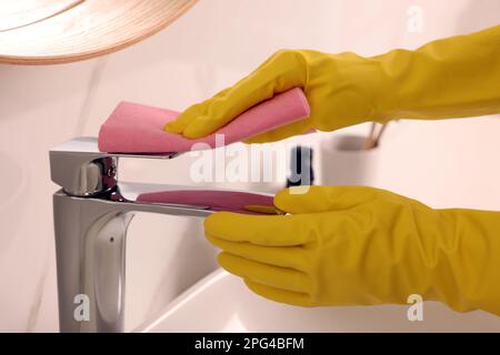 Frau in Handschuhen, die den Wasserhahn des Waschbeckens mit Lappen reinigt Stockfoto