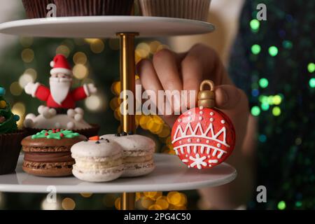 Eine Frau, die Weihnachtsmakaron aus dem Stand nimmt, gegen verschwommene festliche Lichter, Nahaufnahme Stockfoto