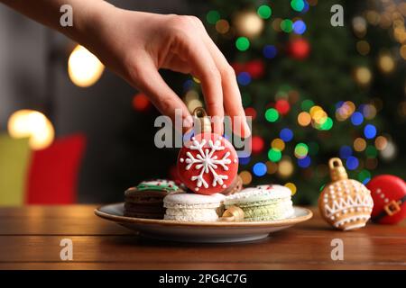 Eine Frau, die Weihnachtsmakarons vom Teller am Holztisch nimmt, vor verschwommenen festlichen Lichtern, Nahaufnahme Stockfoto