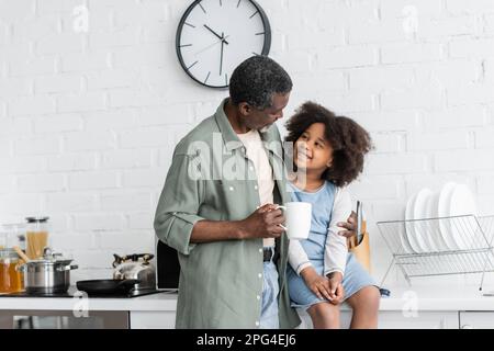 Bärtiger afroamerikanischer Großvater, der eine Tasse Kaffee hält und ein glückliches Kind in der Küche umarmt, Stockbild Stockfoto