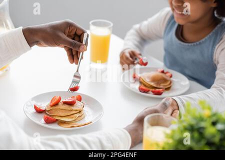 Kurzer Blick auf ein fröhliches afroamerikanisches Mädchen, das beim Frühstück Händchen mit dem Vater hält, Stockbild Stockfoto