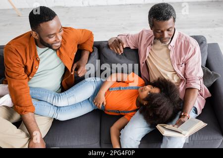 Blick aus der Vogelperspektive auf ein afroamerikanisches Mädchen, das in der Nähe von Vater und Großvater liegt, mit einem Buch zu Hause, ein Bild von der Börse Stockfoto