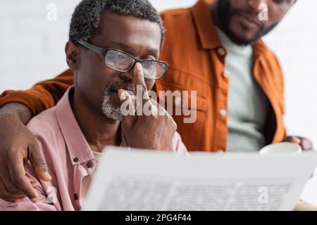Reifer afroamerikanischer Mann mit Brille liest Zeitung in der Nähe eines verschwommenen Sohnes zu Hause, Börsenbild Stockfoto