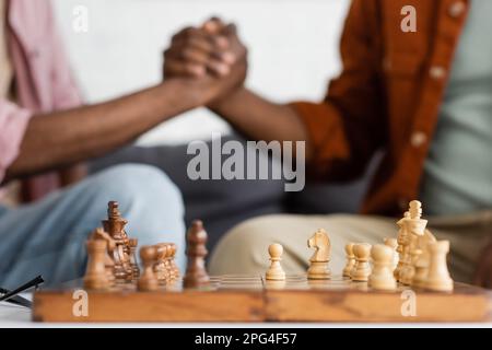 Abgeschnittener Blick auf das Schach an Bord in der Nähe verschwommener afroamerikanischer Vater und Sohn, die zu Hause Händchen halten, Stockbild Stockfoto