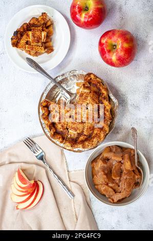 Ein frisch gebackener Apfelkuchen wird auf einem Holztisch serviert, umgeben von einem Teller mit frisch gepflückten roten Äpfeln und einer Tasse Tee Stockfoto