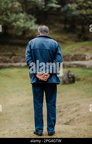 Ein älterer Mann, der auf einer Wiese steht, mit den Händen auf dem Rücken und in die Ferne blickt Stockfoto