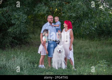Porträt einer hellen, ungewöhnlichen, vielfältigen Familie in der Natur mit dem Samoyerten Hund. Reisen mit Haustieren. Kreative Leute schauen in die Kamera. Grüner botanischer Bac Stockfoto