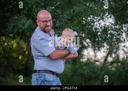 Ein Glatzkopf mit Brille wirft das Kind in die Luft. Vater in Jeans spielt, umarmt sich mit Sohn in der Natur außerhalb der Stadt. Der kleine Junge lacht, amüsiert sich Stockfoto