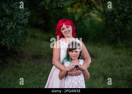 Leuchtend, vielfältig, außergewöhnliche Mutter und Tochter, die sich im Wald umarmen. Die Frau hat rosa Haare, das Mädchen hat grüne Haare. Zärtliche Mutterschaft, glückliches Chi Stockfoto