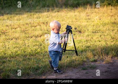 Ein kleiner Junge, der herumspielt, mit einem Stativ als Kamera spielt, auf einer grünen Wiese im Park. Zukünftiger Fotograf. Hobbys für Kinder. Natur bo Stockfoto