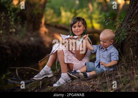 Mädchen und Junge sitzen am Ufer des Flusses und werfen das Origami-Boot aus weißem Papier ins Wasser. Eine Teenager-Schwester mit grünem Haar umarmt den kleinen Bruder. Stockfoto