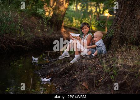 Mädchen und Junge sitzen am Ufer des Flusses und werfen das Origami-Boot aus weißem Papier ins Wasser. Eine Teenager-Schwester mit grünem Haar umarmt den kleinen Bruder. Stockfoto