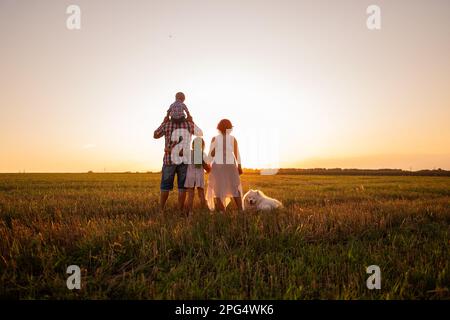Silhouette der Familie mit Samoyerten Hund bei Sonnenuntergang. Gesichtslos, Leute stehen mit dem Rücken zur Kamera. Das Konzept des Reisens, der Freiheit, des Vertrauens in die Fut Stockfoto