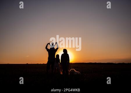 Silhouette der Familie mit Samoyerten Hund bei Sonnenuntergang. Gesichtslos, Leute stehen mit dem Rücken zur Kamera. Das Konzept des Reisens, der Freiheit, des Vertrauens in die Fut Stockfoto