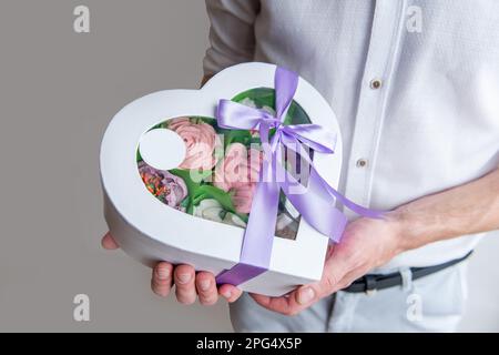 Ein gesichtsloser Mann hält die Form eines Herzens aus süßen Marshmallow-Blumen in den Händen. Geschenk mit lila Band. Zarte, zuckerfreie Rosen auf hellem Hintergrund. Sm Stockfoto