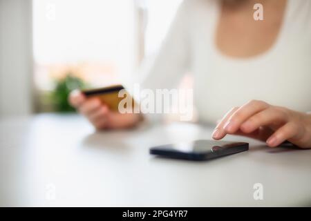 Frau, die online mit Debitkarte und Handy einkauft. Weibliche Hände mit Kreditkarte über einem Handy, das an einem Schreibtisch sitzt und online Waren kauft Stockfoto