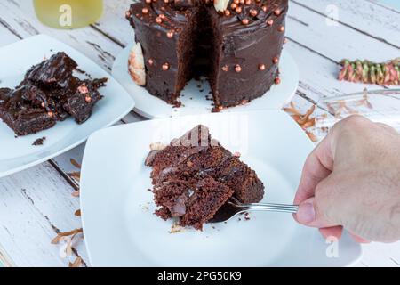 Männliche Hand, die ein Stück Schokoladenkuchen mit bitterem Ganache aufnimmt, gefüllt mit einer Gabel. Stockfoto
