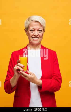 Glückliche Seniorin in rotem Blazer, die ein Glas frischen Orangensaft hält und mit einem Lächeln auf gelbem Hintergrund in die Kamera schaut Stockfoto