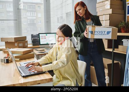 Eine Freiwillige trägt eine Box, während sie an ihrem beschäftigten Kollegen vorbeigeht, der vor dem Computer sitzt und mit dem Handy spricht Stockfoto