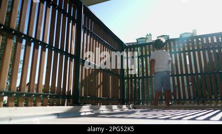 Ein Kind, das am Balkonzaun steht und vom zweiten Stock aus nach draußen blickt, mit Sonnenlicht. Kindersicherung in Holzstangen mit Blick nach draußen Stockfoto