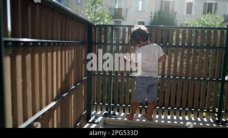 Ein verspielter kleiner Junge stand im Sonnenlicht auf dem Balkon der Wohnung. Kindersicherung auf Terrassenzaun mit Lichtreflexion. Ein Kind schaut nach draußen durch ein hölzernes P Stockfoto