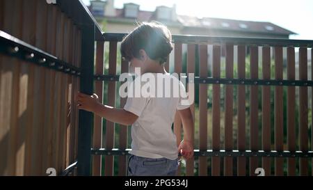 Ein verspielter kleiner Junge stand im Sonnenlicht auf dem Balkon der Wohnung. Kindersicherung auf Terrassenzaun mit Lichtreflexion. Ein Kind schaut nach draußen durch ein hölzernes P Stockfoto