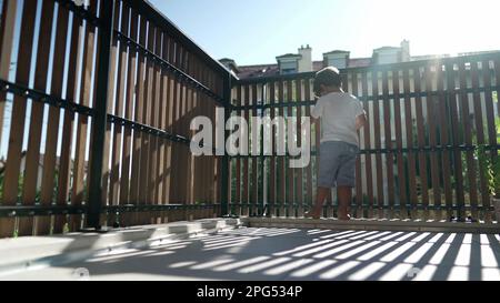 Ein kleiner Junge stand am Balkonzaun und starrte aus dem zweiten Stock heraus mit Sonnenlicht. Kindersicherung in Holzstangen mit Blick nach draußen Stockfoto