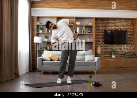 Porträt eines aktiven indianers, der im gemütlichen Wohnzimmer Seitenneigungen mit den Händen an der Hüfte macht. Ein bärtiger junger Mann in Sportkleidung, der regelmäßig Sport macht. Stockfoto