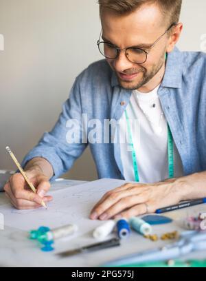 Ein DIY-Designer zeichnet eine Skizze für ein neues Kostümprojekt. Ein selbstlernender Schneider mit Brille arbeitet am Tisch. Ein lächelnder Mann in einem blauen Hemd Stockfoto