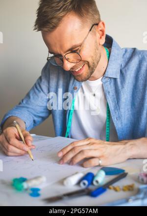 Ein Schneider mit Brille. Ein lächelnder Mann in einem blauen Hemd macht eine Skizze für das Nähen von Kleidung als Hobby. Ein DIY-Designer zeichnet eine Skizze für ein neues Kostüm Stockfoto