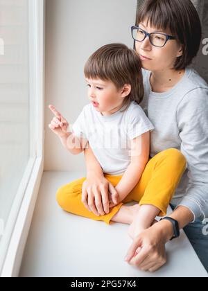 Traurige Frau schaut ins Fenster. Zwangsaufenthalt zu Hause wegen COVID-19-Quarantänepandemie. Mutter und Sohn. Mutter und Kind. Stockfoto