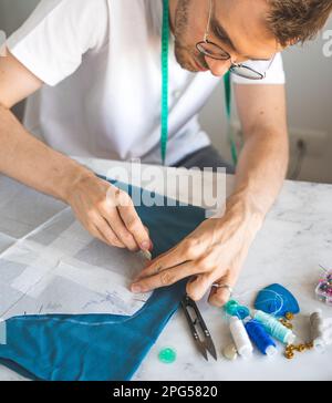 Ein männlicher Heimwerker überträgt eine Vorlage in ein Stoffstudio. Ein selbstlernender Schneider in einem weißen T-Shirt und einer Brille arbeitet mit Stoff und Muster Stockfoto
