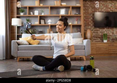 Sportlicher afroamerikanischer Lehrer mit modernen Geräten, um morgens mit dem Heimtraining zu beginnen. Attraktive junge Frau in Sportbekleidung hört Followern zu, während sie auf einer Yoga-Matte sitzt. Stockfoto