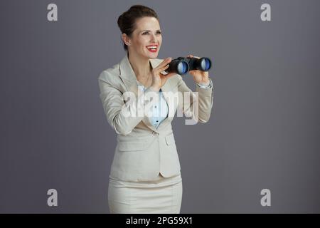 Eine glückliche trendige Geschäftsfrau mittleren Alters in einem leichten Business-Anzug mit Fernglas isoliert auf grauem Hintergrund. Stockfoto