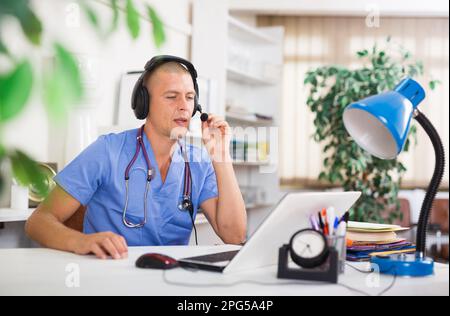 Arzt in Ohrhörern mit Online-Konsultation mit dem Patienten auf dem Laptop Stockfoto