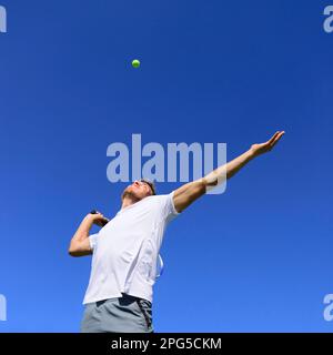 Ein Tennisspieler, der in Position ist, um den Ball in die Luft zu schlagen. Tennis im Freien im Sommer mit blauem Himmel. Aktive Sportler machen Stockfoto