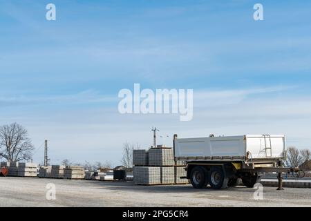 Baustelle mit leerem Pkw-Anhänger und Baumaterial auf Paletten. Blauer Himmel mit hellweißen Wolken. Stockfoto