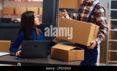 Junger Mann analysiert Kisten mit Verbrauchsmaterial im Lager, um weibliche Mitarbeiterin bei der Finanzplanung und Geschäftsentwicklung zu unterstützen. Team von Kollegen, die an Lagerbestand und Versand arbeiten. Handgeführte Aufnahme. Stockfoto