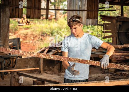 Junger Mann, der in einem Sägewerk arbeitet und Schutzhandschuhe trägt. Junger Teenager mit Holzbrettern, frisch geschnitten von einer Maschine. Stockfoto