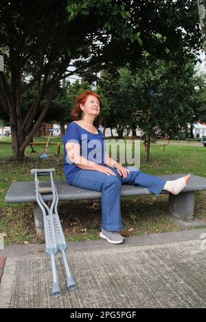Lateinische reife Frau mit einer Verletzung, die auf einer Parkbank saß, mit einem Verband am linken Fuß und Krücken neben ihr. Konzepte: Positive Atti Stockfoto