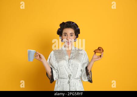 Aufgeregte junge Frau in Seidenbademantel mit Lockenwicklern zum Frühstück auf orangefarbenem Hintergrund Stockfoto