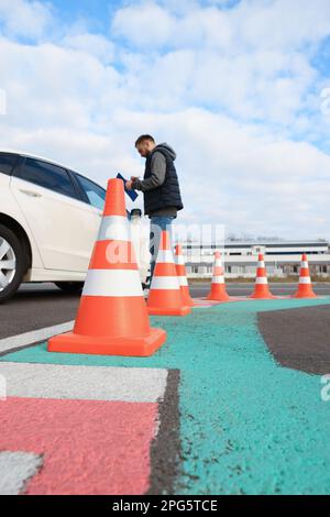 Ausbilder mit Klemmbrett in der Nähe des Fahrzeugs im Freien, konzentrieren Sie sich auf den Verkehrskegel. Fahrschulprüfung Stockfoto