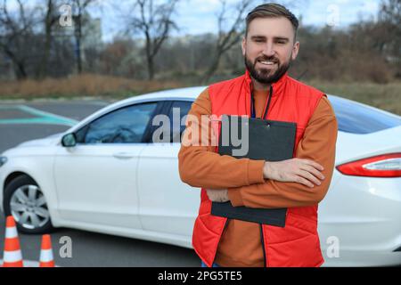 Kursleiter mit Klemmbrett in der Nähe des Fahrzeugs auf der Teststrecke. Fahrschule Stockfoto