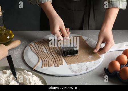 Frau, die Soba (Buchweizennudeln) mit Schneidevorrichtung am grauen Tisch macht, Nahaufnahme Stockfoto