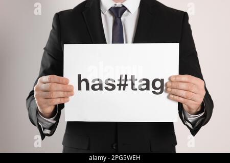 Ein Mann hält ein Blatt Papier mit Wort- und Symbol-Hashtag auf hellem Hintergrund, Nahaufnahme Stockfoto