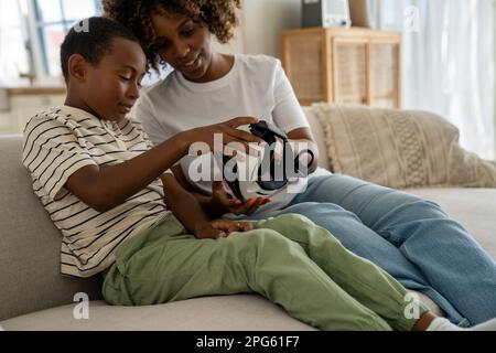Lächelnde Mutter aus gemischter Rasse mit Sohn sitzt auf der Couch und hält das VR-Headset für ein Videospiel Stockfoto